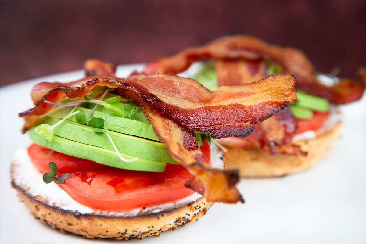 Two bagels with avocado, bacon, and tomatoes on a white plate.