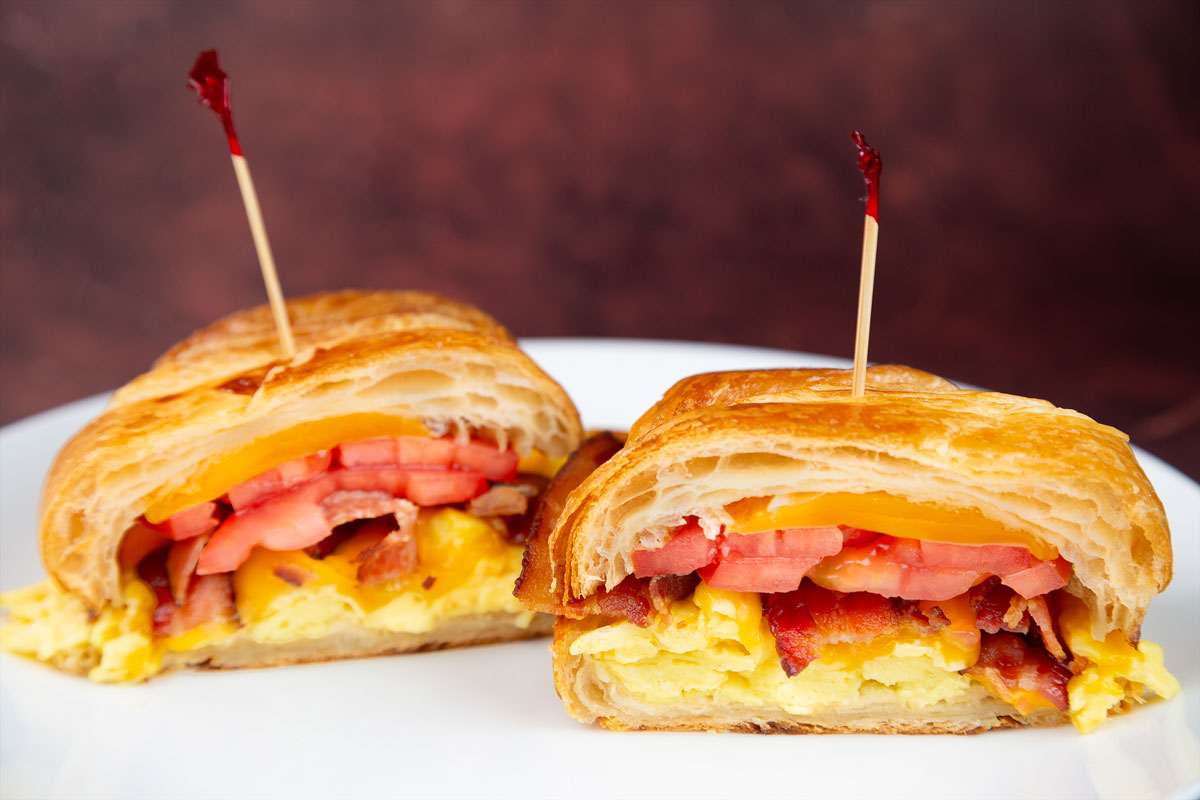 A close-up of a croissant breakfast sandwich with egg, tomato, and bacon served on a white plate.