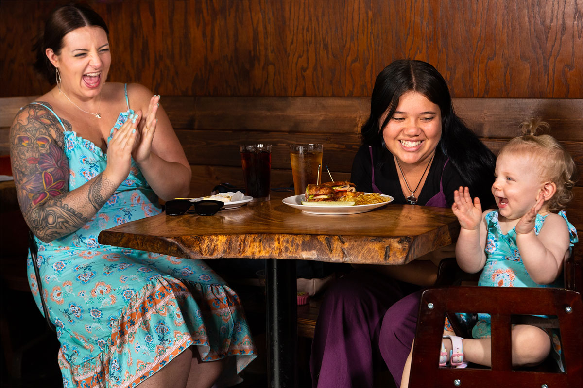 A woman adorned with tattoos who claps and smiles with her baby and a friend in Wailuku Coffee Co. café on Maui.