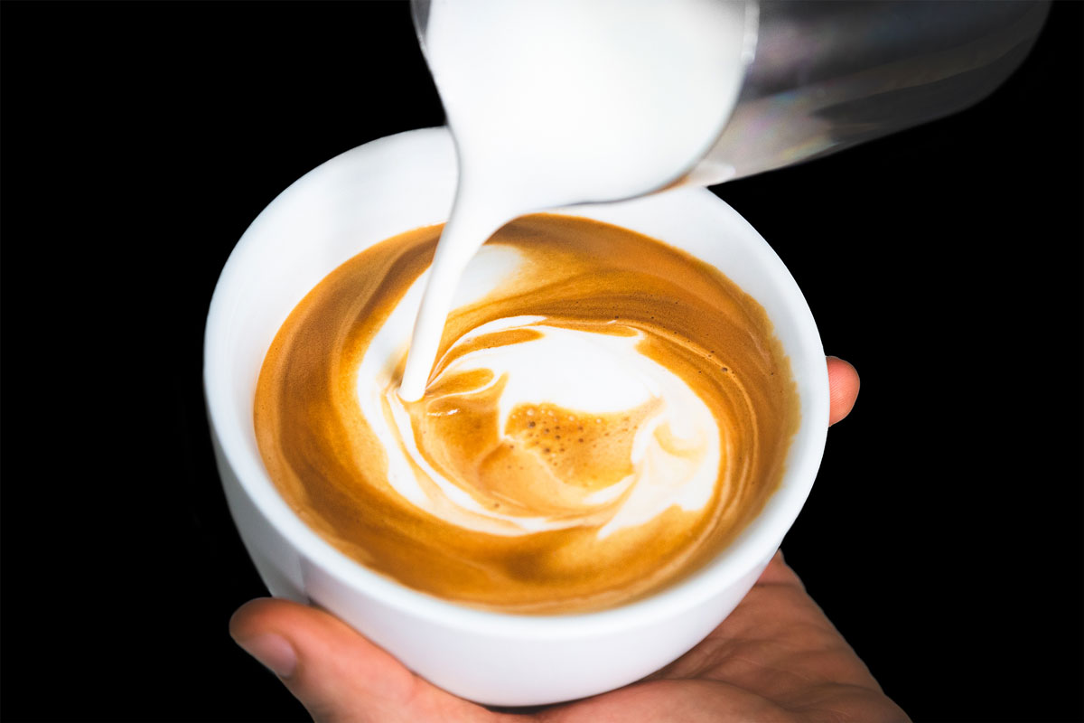 A person pouring a creamy latte into a white cup with a black background.
