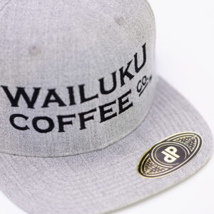 The grey snapback hat features a side view of the Wailuku Coffee Co logo embroidered in black and a limited edition label.